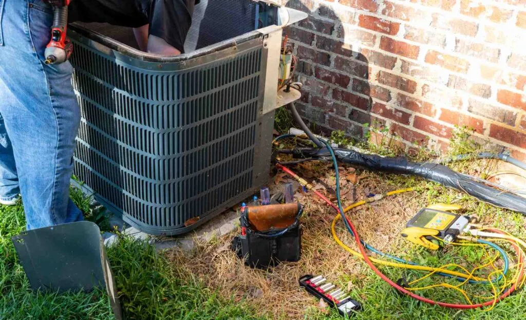 Technician servicing an outdoor condenser unit with tools and gauges during a year-round HVAC maintenance tune-up.